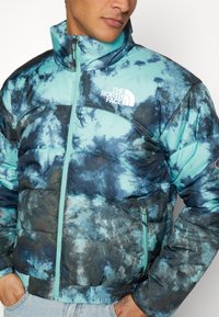 Hombre que lleva una chaqueta acolchada de tie-dye en azul y negro con cuello alto, frente con cremallera, y el logo de The North Face en el lado izquierdo del pecho.