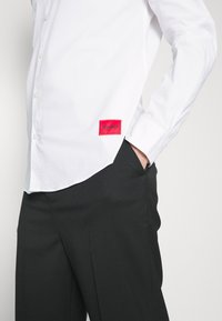 Vit skjorta med knappar och en röd broderad "HUGO"-patch på nedre vänstra delen. Skjortan har långa ärmar och en figurnära design. svarta byxor fulländar stilen.