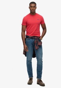 Camiseta de algodón roja con logo, combinada con unos jeans de mezclilla azul. Una camisa de cuadros negra y roja atada a la cintura, botas de cuero marrón.
