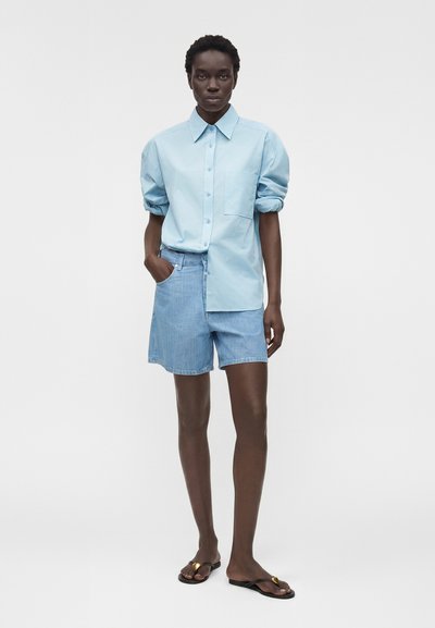 Άτομο στέκεται φορώντας ανοιχτό μπλε πουκάμισο με κουμπιά και μανίκια που είναι τυλιγμένα, ανοιχτό μπλε τζιν σορτς και μαύρα σανδάλια με χρυσές λεπτομέρειες.