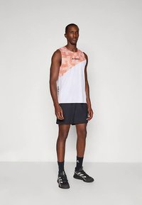 Balta sportinė liemenė su oranžinių dryžių dizainu, derinama su juodais šortais. Pagaminta iš kvėpuojančios medžiagos ir turi logotipo akcentus. Juodi bateliai.