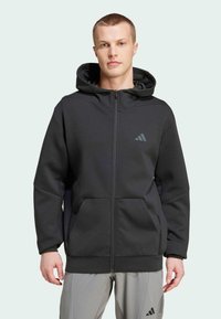 Haut à capuche zippé noir en tissu doux, doté d'une grande capuche, de poches avant et d'un logo subtil sur la poitrine. Ajustement texturé et confortable.