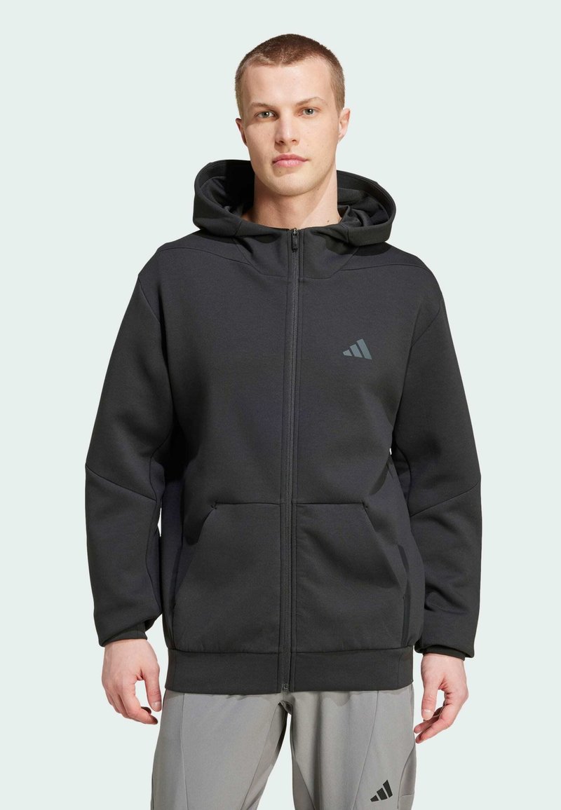 Haut à capuche zippé noir en tissu doux, doté d'une grande capuche, de poches avant et d'un logo subtil sur la poitrine. Ajustement texturé et confortable.