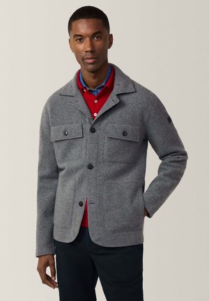 Grijze wollen jas met een knoopsluiting aan de voorkant, twee borstzakken en een geknopt kent. Draagt over een rood shirt met een blauwe kraag. Gladde textuur.