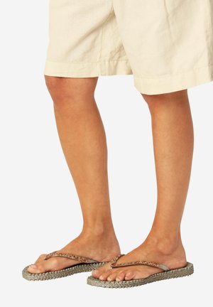 Piernas con pantalones cortos beige y chanclas grises con suelas trenzadas y tiras brillantes en color oro rosa sobre un fondo blanco.