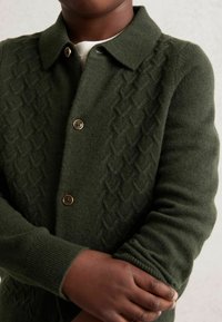 Nahaufnahme einer Person, die einen dunkelgrünen strukturierten Cardigan mit Knöpfen trägt, die Arme verschränkt hat, und ein weißes Hemd sichtbar ist, das unter dem Kragen hervorblitzt.