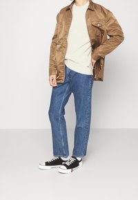 Braune Satinjacke, leichter, gerippter beiger Pullover, blaue Straight-Leg-Jeans und schwarze Sneakers mit weißen Sohlen und roten Akzenten.