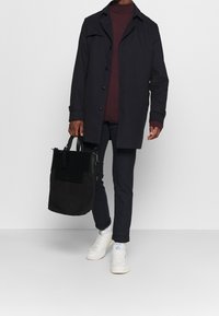 Schwarzer Mantel mit Knopfleiste, burgunderroter Rollkragenpullover, dunkle Hose und weiße Turnschuhe. Trägt eine schwarze Tragetasche mit zwei Griffen.