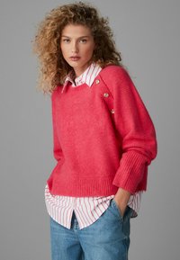 Lyserød strikket sweater med ribbet kant og dekorative knapper, lagdelt over en hvid og lyserød stribet skjorte med krave, parret med blå jeans.