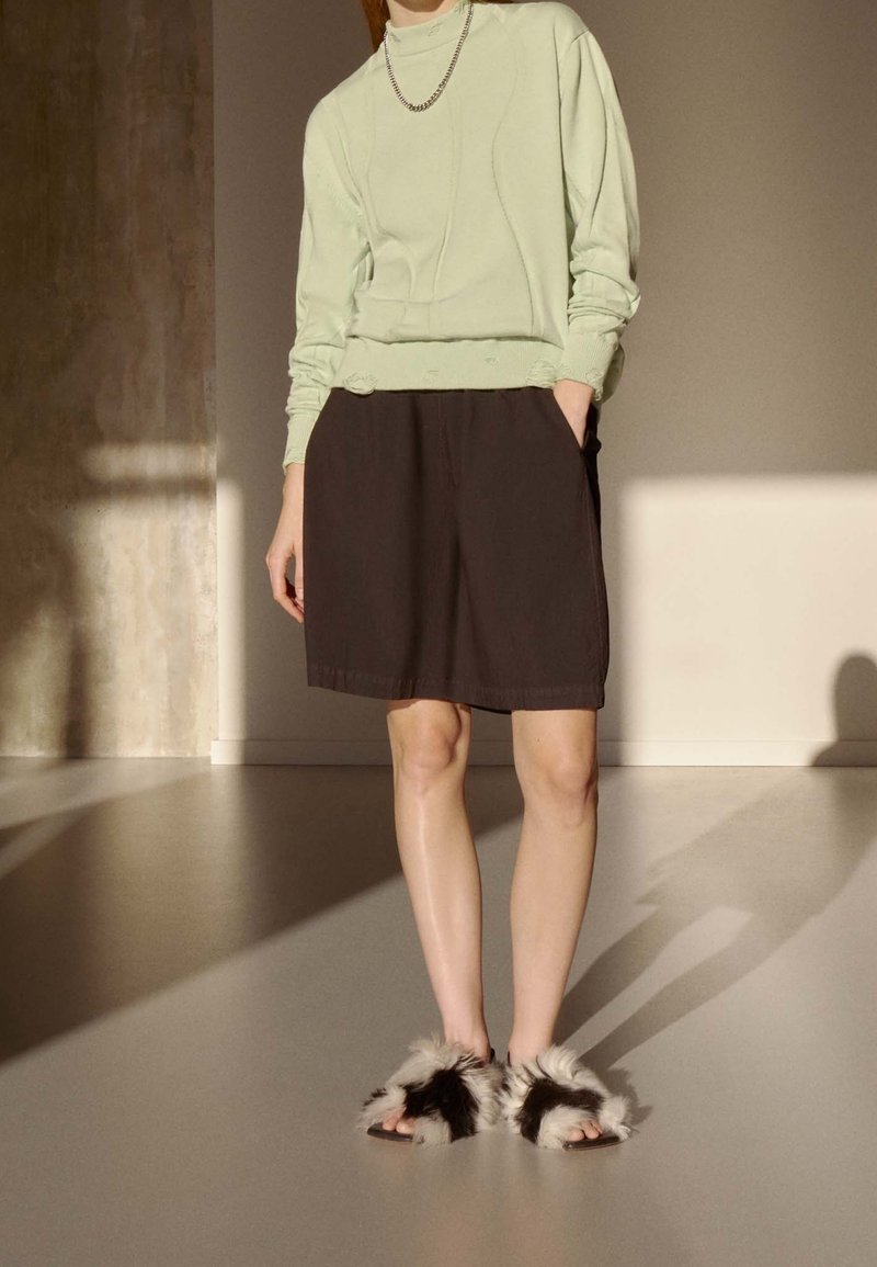 Ljusgrön sweatshirt med ribbad nederkant, i kombination med mörkbruna shorts och sandaler i svart och vitt fuskpäls. Inomhusmiljö med mjukt ljus.