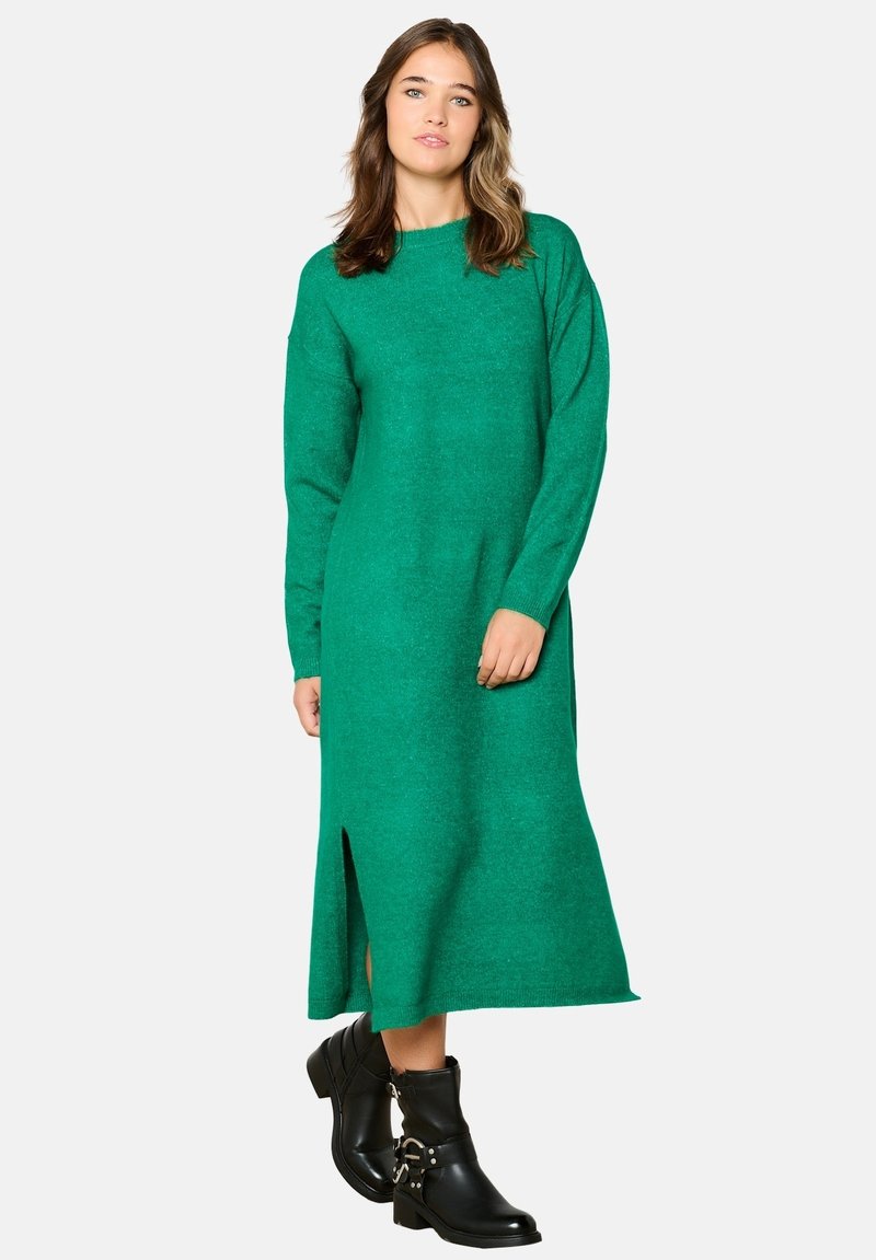 Abito verde lavorato a maglia con maniche lunghe, scollatura alta e spacco laterale. Abbinato a stivaletti neri alla caviglia con dettagli in metallo a forma di fibbia.