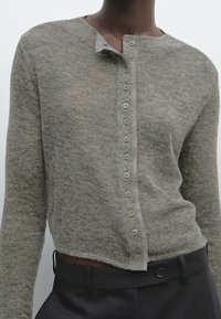 Henkilö, jolla on päällään vaalea harmaa napillinen villatakki, jossa on pieniä nappeja, ja tummanharmaat korkeat housut.