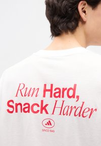 Άτομο που φοράει λευκό πουκάμισο με κόκκινο κείμενο «Τρέξε Σκληρά, Σνακάρεις Περισσότερο» και ένα μικρό λογότυπο με την ένδειξη «Από το 1949» στην πλάτη.