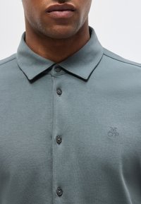 Άνδρας που φοράει μπλε-γκρι πουκάμισο με γιακά και κουμπιά, με μικρό λογότυπο "MOP" κεντημένο στο στήθος, εμφανιζόμενο από τον λαιμό μέχρι το μέσο του κορμού.