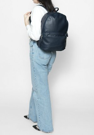 Sac à dos en cuir navy avec une texture lisse, poche zippée et bretelles réglables, présenté sur une personne portant un jean bleu.