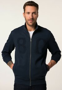 Sudadera azul marino con cremallera y diseño elevado "80" en el frente. Cuenta con puños y dobladillo acanalados, bolsillos laterales y cuello alto. Textura de tela suave.