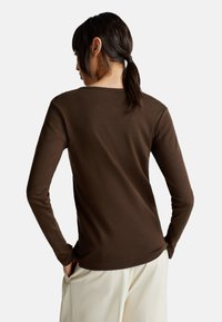 Vrouw met donker haar in een paardenstaart, gekleed in een aansluitend bruin shirt met lange mouwen en lichtbeige broeken, staand met de handen in de zakken, met de rug naar de kijker gekeerd.