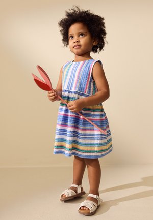 Jeune enfant aux cheveux bouclés portant une robe rayée colorée et des sandales blanches, tenant une grande fleur rouge en accessoire sur un fond uni.