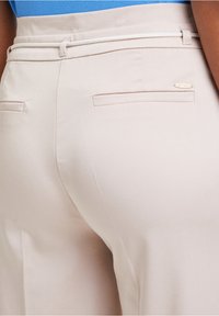 Beige hoge taille broeken met een gladde textuur, voorzien van twee achterzakken en een witte trekkoord aan de tailleband. Subtiele merkdetails aanwezig.
