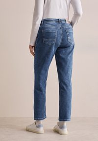 Jean bleu foncé avec une finition légèrement délavée, cinq poches et un design à jambe droite, porté avec des baskets blanches et des chaussettes légères.
