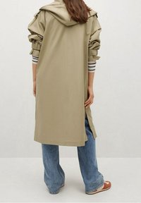 Trench-coat kaki avec capuche, manches froncées et fentes latérales, associé à un jean large bleu clair et des sandales marron.