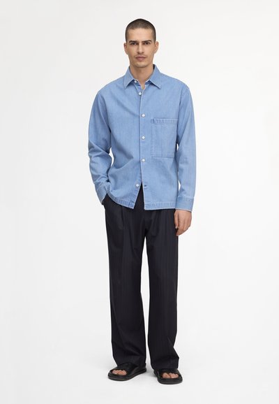 Homme debout portant une chemise bleu clair boutonnée, un pantalon noir ample et des sandales noires à bouts ouverts sur un fond blanc uni.