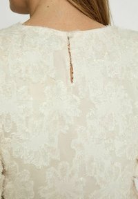 Textured cream fabric with floral pattern, featuring a keyhole detail at the back and frayed edges along the neckline.