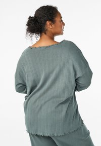 Loose-fitting, long-sleeve top in muted teal fabric with a textured rib pattern, featuring a raw, scalloped neckline and hem.