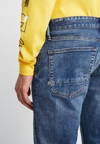 Blå jeans i denim med en blektvättad finish, med en bakficka som har stickningsdetaljer. Matchas med en gul långärmad tröja.