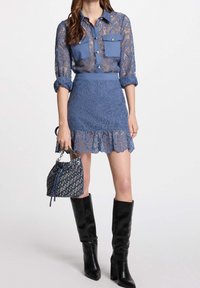 Blouse en dentelle bleue avec deux poches, associée à une jupe en dentelle assortie avec un ourlet à volants. Bottes noires montantes jusqu'aux genoux et mini-sac à motif.