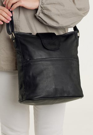 Person holding black leather shoulder bag with flap closure, wearing beige long-sleeve top and white pants against plain background.