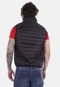 Gilet imbottito nero con colletto alto e design a righe orizzontali, indossato sopra una camicia rossa a maniche corte. Presenta una superficie liscia e lucida.