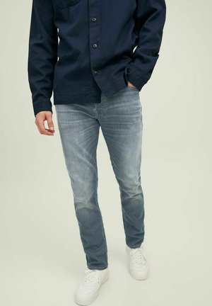 Slim-fit light blue denim jeans with a faded finish, paired with a navy button-up shirt and white sneakers. Simple design and classic style.