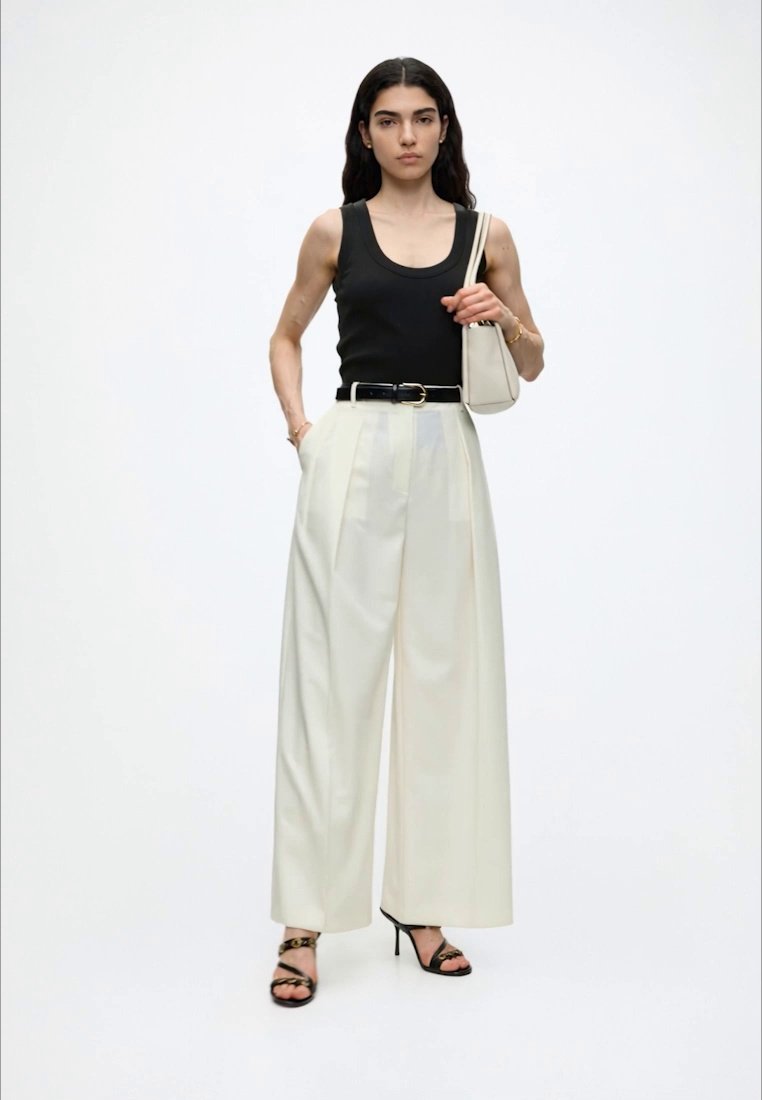 Femme debout portant un haut noir sans manches, un pantalon blanc large, une ceinture noire, des talons noirs à brides et tenant un sac blanc à l'épaule.
