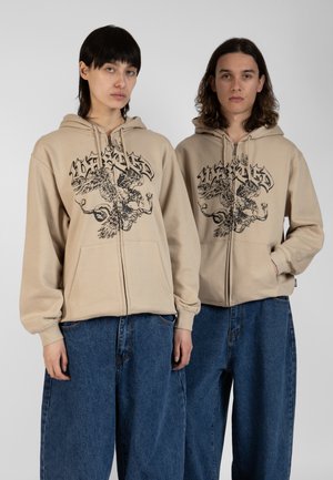 Beige zip-up hoodie featuring a black graphic design of a mythical creature, with large front pockets and elasticized cuffs. Paired with loose denim.