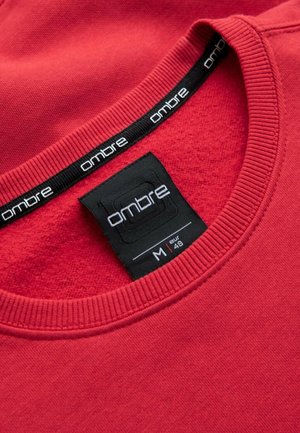 Sweatshirt rouge avec col côtelé, présentant une étiquette noire avec le mot "ombre" en texte blanc. Taille indiquée M (48 EUR). Tissu doux et texturé.