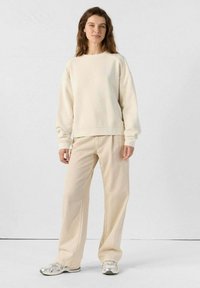 Felpa beige dal taglio ampio e con collo rotondo, abbinata a pantaloni beige a gamba larga coordinati. Il modello indossa sneakers argentate per un contrasto.