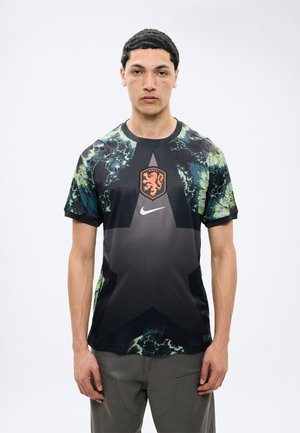 Νέος άνδρας στέκεται με το βλέμμα προς τα εμπρός, φορώντας μια μαύρη αθλητική φανέλα με πράσινα αφηρημένα μοτίβα και ένα πορτοκαλί έμβλημα λιονταριού στο στήθος.