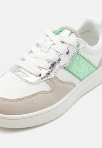 Basket blanche avec des accents verts et argent métallisé, des détails en suède et une finition texturée. Présente un système de laçage et des sections perforées.
