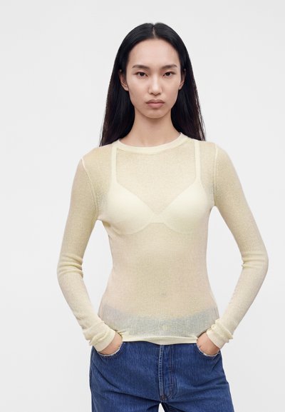 Jeune femme aux longs cheveux noirs porte un top crème transparent à manches longues sur un soutien-gorge crème, associé à un jean bleu, les mains dans les poches.