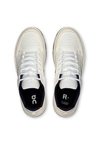 Baskets blanches de sport avec un dessus perforé, des lacets plats et une semelle noire. Les caractéristiques comprennent un texte de logo à l'intérieur et une couture distinctive.