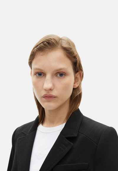 Blazer noir avec un design structuré et un tissu texturé, associé à une chemise blanche à côtes. Le col a des bords nets et un minimum de détails.