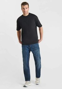 Maglietta nera a maniche corte abbinata a jeans blu con vestibilità rilassata, variazioni di texture e dettagli di cucitura sottili. Indossata con scarpe da ginnastica chiare.