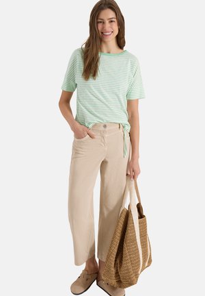 Young woman smiling, wearing a light green striped shirt, beige wide-leg pants, beige slip-on shoes, holding a large woven tote bag.