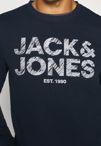 Marinblå sweatshirt med stor, sliten vit text "JACK & JONES" och "EST. 1990" centrerad på framsidan. Tillverkad av bomullsmaterial, med rund hals.