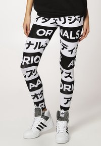 Legíny s černobílými bloky s nápisem "ORIGINALS" a japonskými znaky, nošeny s bílými vysokými teniskami Adidas.