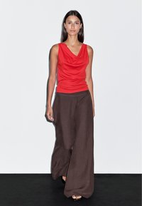 Débardeur rouge à col cowl associé à un pantalon large marron foncé. Le tissu a une texture lisse, avec une coupe décontractée et un design simple.