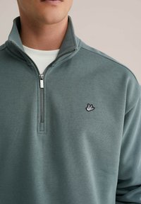 Svetlo zelena jopa na zadrgo iz mehke tkanine, z majhnim črnim logotipom ptice na levi strani prsnega koša in visokim ovratnikom s srebrno zadrgo.