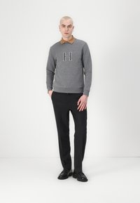 Grauer Pullover mit braunem Kragen, ausgestattet mit einem großen "H"-Logo. Kombiniert mit schwarzen, taillierten Hosen und dunklen Schuhen.