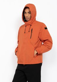 Oranje waterdichte jas met capuchon, elastische manchetten, rits aan de voorkant en zijzakken. Heeft zwarte ritsaccenten en een logo op de mouw.